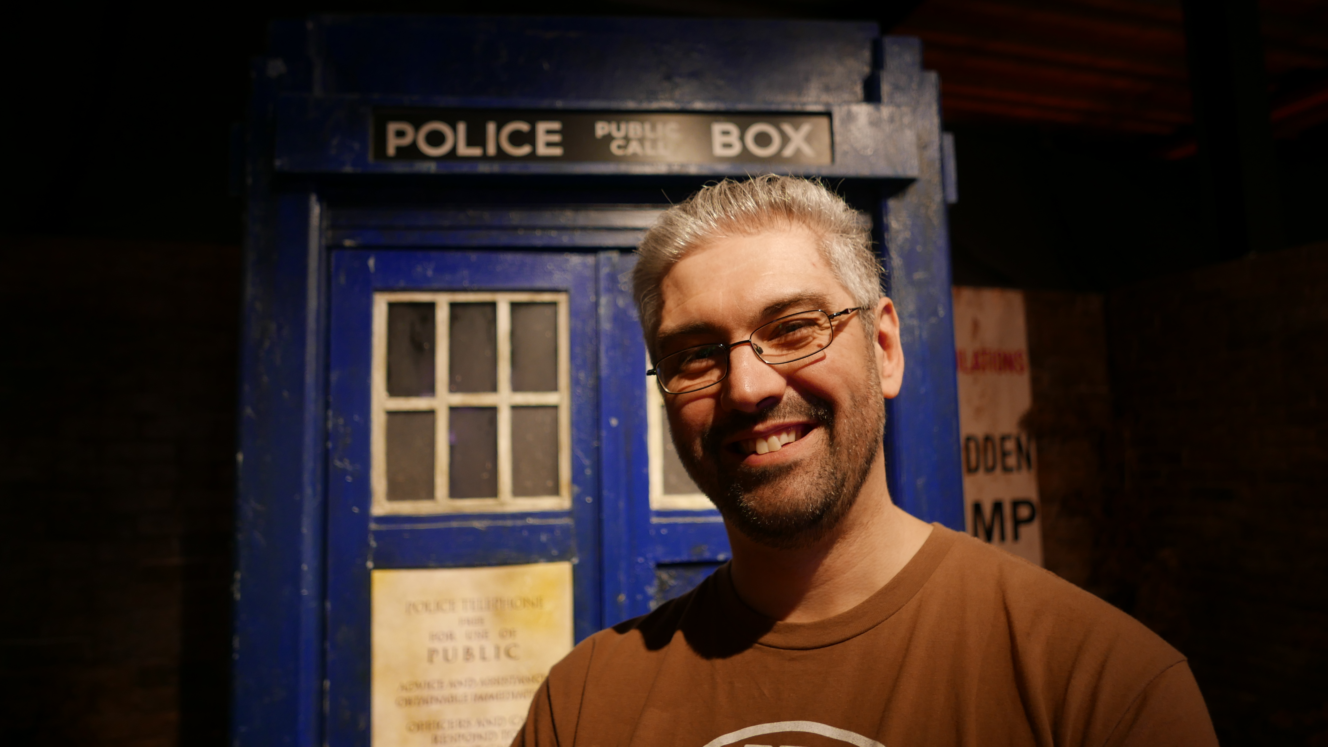 A smiling bearded man with glasses, Si Hart, is standing in front of the TARDIS
